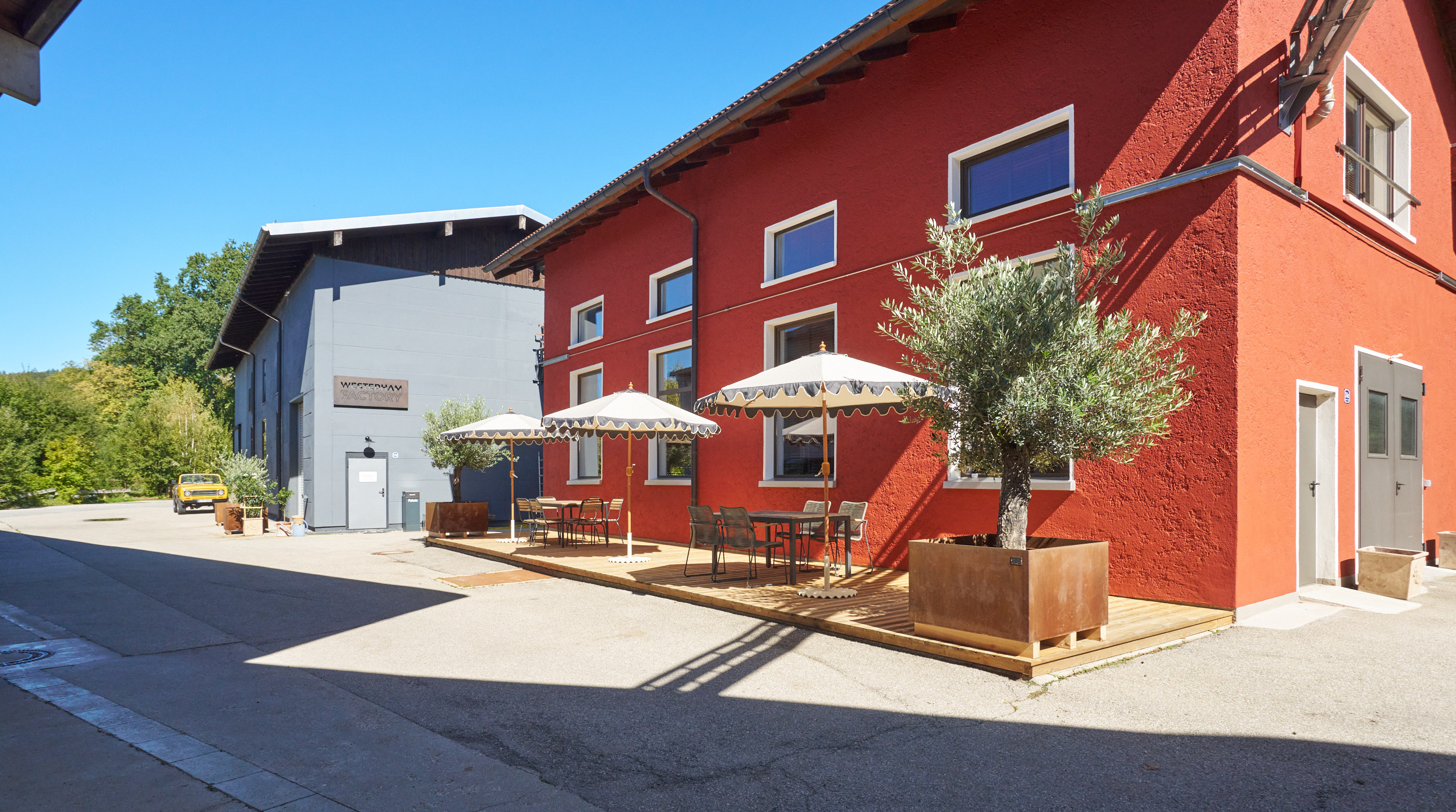 Außenansicht der Westerham Factory mit rotem Gebäude, Holzterrasse und Sonnenschirmen